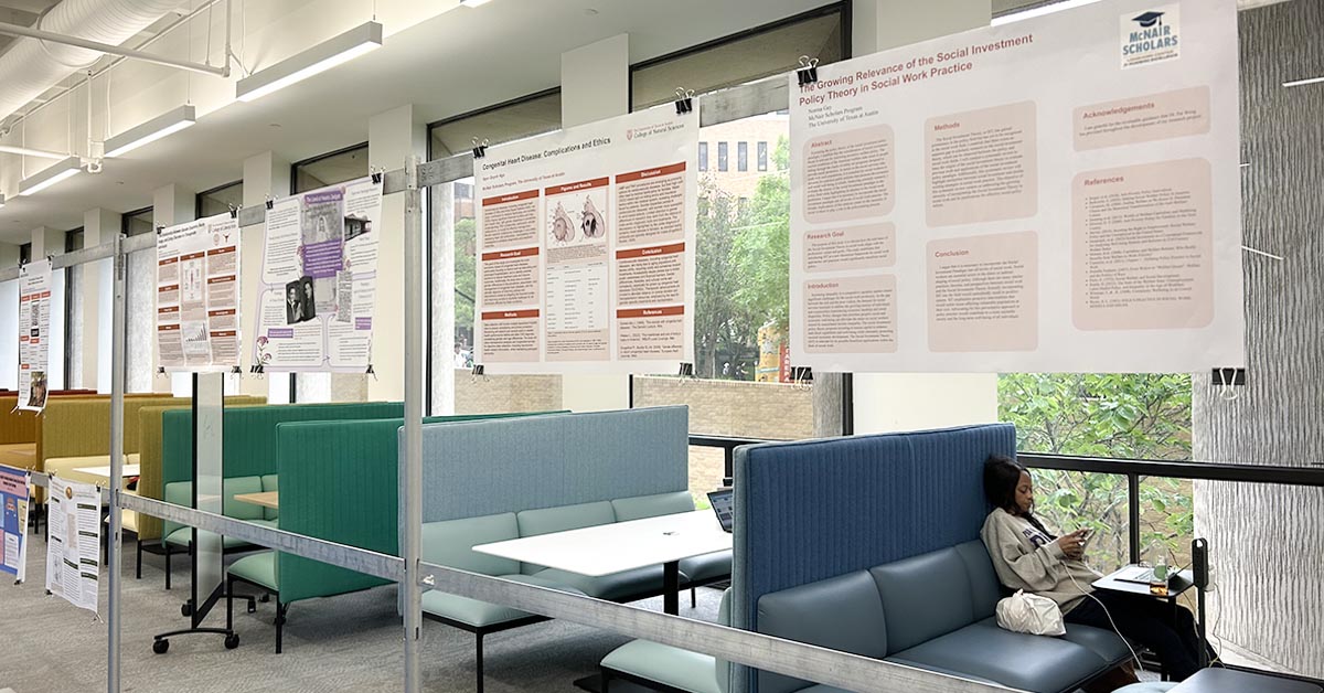 Research posters hanging on free-standing longitudinal frame in study area of scholars lab. Cafe benches in bright colors with solitary student studying and windows with a view of outdoors in the background.